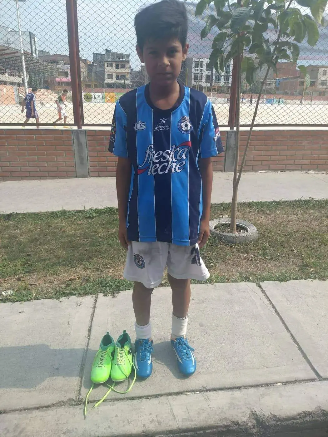 Young soccer player stands with green cleats beside him on sidewalk.